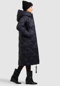 Sieviete, valkājot garu segtu tumši zilu mēteļa ar kapuci, melnu cepuri, melnas zeķubikses un melnus kāju iešļūcenes ar platformu, skatās sāniski ar rokām kabatās.