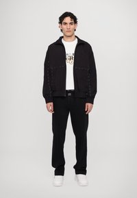 Schwarze Jacke mit kontrastierenden weißen Nähten, getragen über einem weißen T-Shirt mit grafischem Text, kombiniert mit schwarzen Hosen und weißen Schuhen.