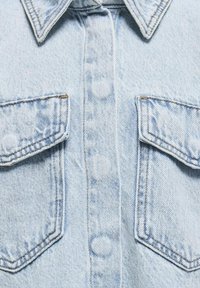 Ljusblå denimskjorta med spetsig krage, två bröstfickor och knäppningar. Tyget har en något blek textur.