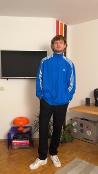 Chaqueta de chándal azul con cierre y rayas blancas, pantalones negros y zapatillas blancas. Con un fondo de sala de estar moderna.