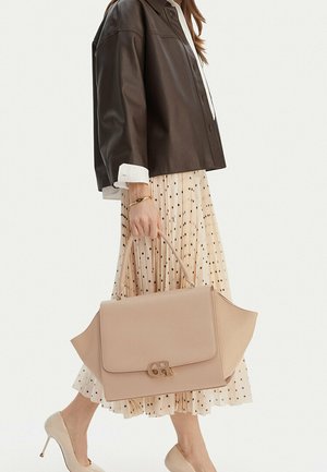 Donna con gonna plissettata a pois, giacca marrone scuro, tacchi beige e che tiene una grande borsa beige con logo dorato sul manico.