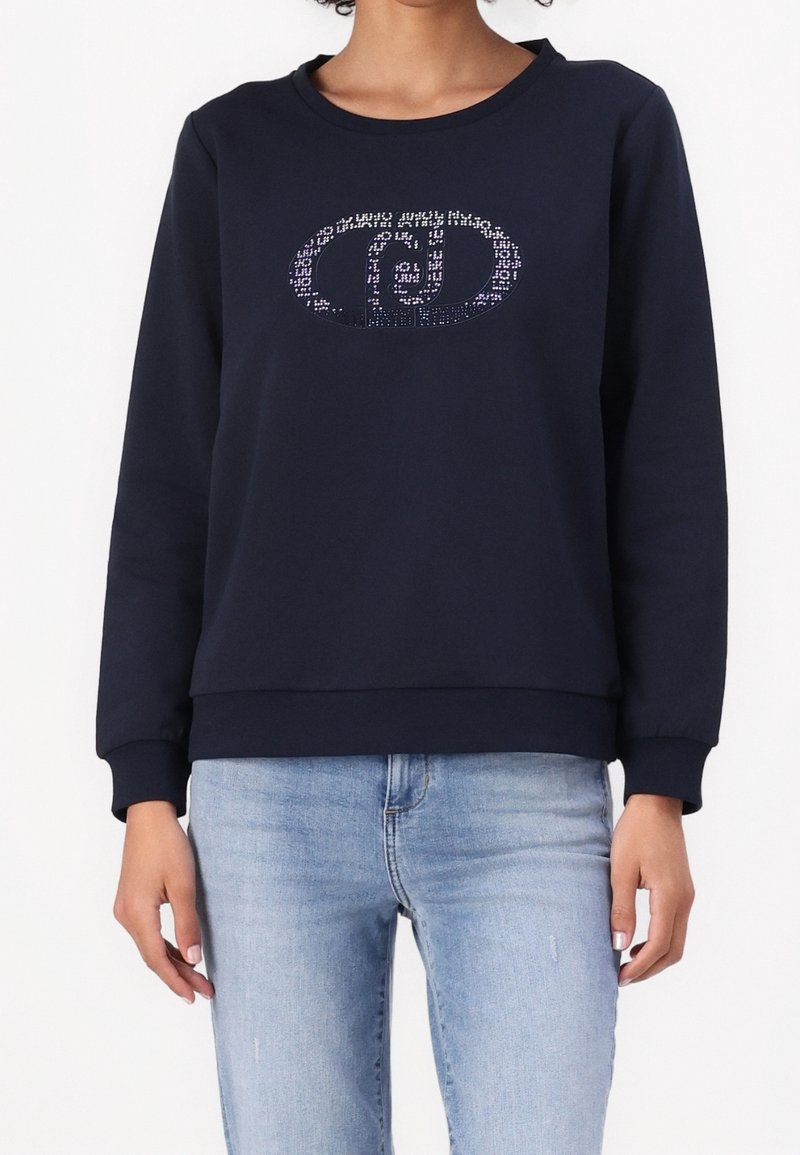 Marineblauwe sweatshirt met lange mouwen, geribbelde manchetten en zoom, voorzien van een sprankelende ovale logo aan de voorkant. Gekleed met lichtblauwe jeans.