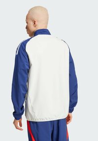 Chaqueta cortavientos blanca y azul marino que cuenta con mangas largas, un cuello alto y paneles laterales en color azul que contrastan. Puños y dobladillo elásticos.