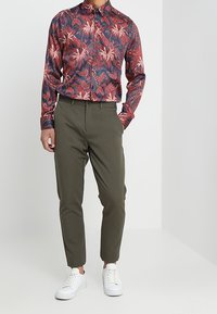 Chemise homme à manches longues en bleu marine avec un motif de feuilles rouges et blanches, associée à un pantalon kaki fuselé et des baskets blanches.