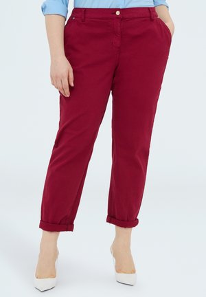 Kobieta w podwiniętych bordowych spodniach, jasnoniebieskiej koszuli i białych szpilkach, stojąca z jedną ręką w kieszeni.