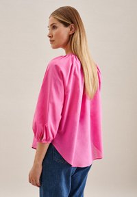Helle pinke Bluse mit lockerer Passform, ausgestattet mit Puffärmeln, einem gerafften Halsausschnitt und einem hohen, tiefen Saum, kombiniert mit blauen Jeans.