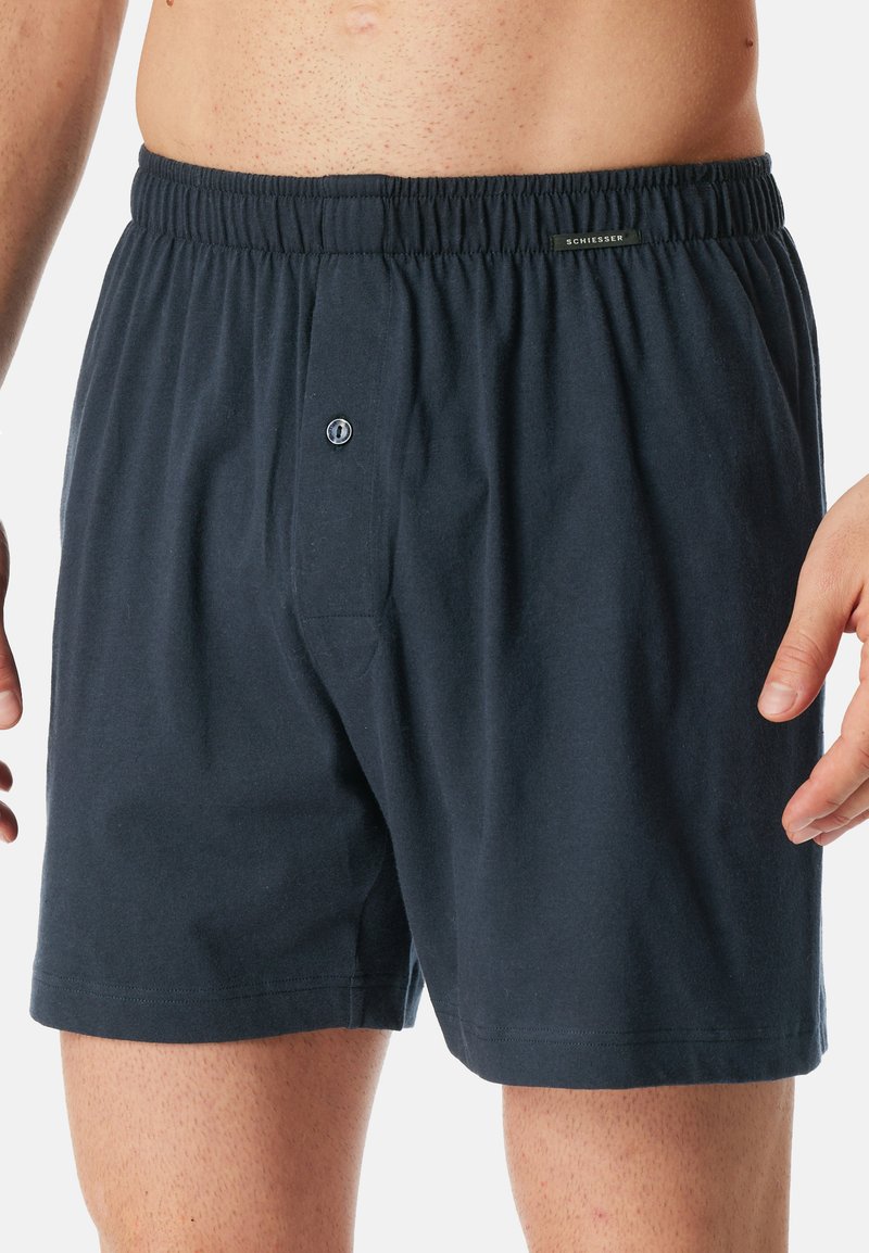 Boxer in cotone blu navy scuro da uomo con vita elastica, patta con un singolo bottone e texture morbida; dettagli di branding minimalisti visibili.