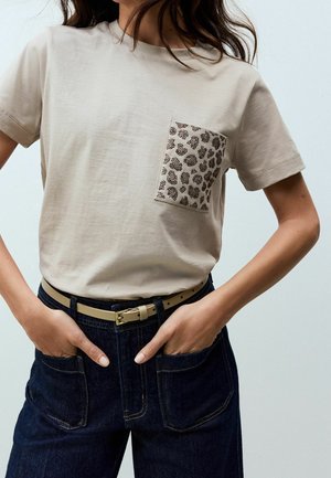 Beżowa koszulka z kieszonką w panterkę, wpuszczona w ciemnoniebieskie dżinsy z beżowym paskiem, ręce modela w przednich kieszeniach.