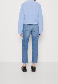 Žena stojící zády, oblečená v lehké modré strukturované svetru, rovné modré džíny a černé platformové boty na jednobarevném pozadí.