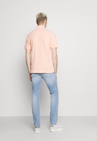 Helles rosa Poloshirt mit klassischem Kragen, kurzen Ärmeln und lockerer Passform, kombiniert mit hellen Jeans und weißen Sneakers.