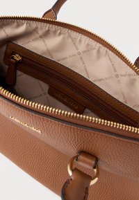 Καφέ δερμάτινη γυναικεία τσάντα με φερμουάρ, διακοσμημένη με μπεζ επένδυση, εσωτερική τσέπη και μεταλλικές λεπτομέρειες.