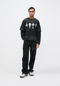 Črna prevelika majica s potiski v beli barvi, kombinirana z ohlapnimi črnimi hlačami z srebrnimi poudarki in stranskimi okraski.