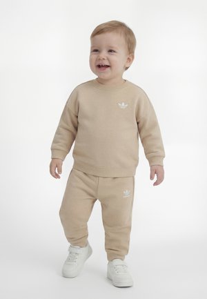 Småbarn står og smiler, iført beige Adidas sweatshirt og bukser med hvide sneakers, på en ensfarvet hvid baggrund.