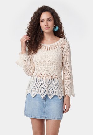 Donna con capelli ricci indossa grandi orecchini blu, un maglione di cotone crema lavorato a uncinetto con orlo smerlato e una gonna di jeans azzurro chiaro su sfondo grigio.