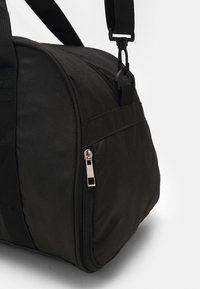 Zwarte duffel tas van textuurstof, met een afneembare schouderriem, zijrits en een gebogen vorm. Voorzien van een zilveren metalen ritstrekkertje.