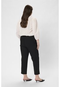 Mujer de pie, de espaldas, vistiendo una blusa blanca transparente, pantalones negros de cintura alta, zapatos de tacón bajo negros y pulseras de perlas en la muñeca derecha.