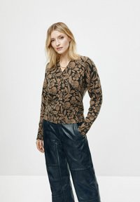 Sweter w kwiatowy wzór w kolorze czarnym z beżowymi detalami, z dekoltem w kształcie litery V i długimi rękawami, zestawiony z ciemnymi spodniami przypominającymi skórę.