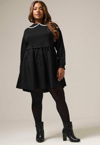 Musta mekko, jossa on pitkät hihat ja istuva yläröijy, joka sisältää kerätty vyötärö ja yksivärinen musta hame. Yhdistetty mustiin korollisiin nilkkasaappaisiin.