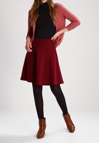 Burgundy A-linje kjol med veck, kombinerad med en svart polokrage och en texturerad rosa kofta, kompletterad med bruna ankelstövlar.