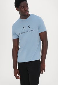 Gaiši zila īsām piedurknēm T-kreklu, kas izgatavots no mīksta auduma, ar melnu uzdrukātu logo "A|X ARMANI EXCHANGE" krūšu daļā.
