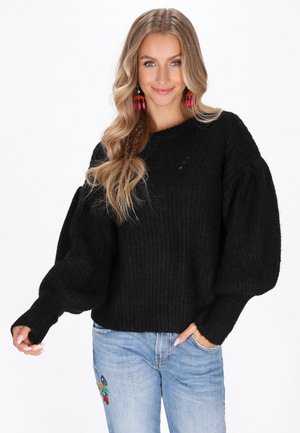 Zwarte gebreide trui met geribbelde textuur, brede mouwen en een ronde hals. Gecombineerd met blauwe jeans met kleurrijke borduursels.