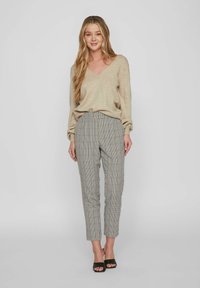 Beiger V-Ausschnitt Pullover, lockere Passform, kombiniert mit grauen karierten Stoffhosen, schmal zulaufendes Bein, Knöchellänge, und schwarzen Absatzsandalen. Glatte Texturen überall.