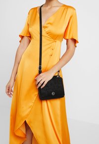 Abito di raso giallo con maniche corte e design avvolgente, abbinato a una piccola borsa a tracolla in finta pelle nera con logo in argento.