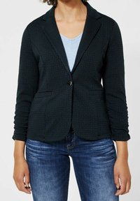 Tmavě zelený blazer vyrobený z texturované tkaniny s jemným vzorem, disponující uzávěrem na jeden knoflík a dvěma předními kapsami. Nosený přes světle modrý top.