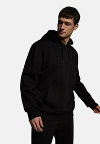 Czarna bluza z kapturem wykonana z miękkiej, teksturowanej tkaniny. Posiada kieszeń kangurzą, regulowany sznurek i ściągacze na rękawach. Prosty, minimalistyczny design.