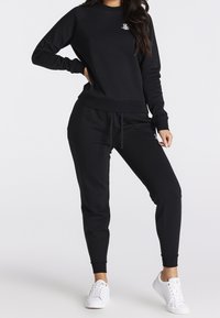 Felpa nera in pile con polsini e fondo a costine, caratterizzata da un piccolo logo bianco. Abbinata a pantaloni jogging neri coordinati e sneakers bianche.