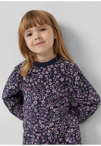 Junges Mädchen mit hellbraunen Haaren, das ein navyblaues Kleid trägt, das mit kleinen lila und weißen Blumen bedeckt ist, steht mit den Händen hinter dem Rücken.