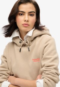 Felpa beige con cappuccio regolabile, hardware nero e logo ricamato rosso. Indossata sopra una camicia bianca con colletto. Texture morbida.