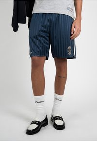 Homme portant un short rayé bleu marine, des chaussettes blanches avec un texte noir et des mocassins noir et blanc, debout devant un fond uni.