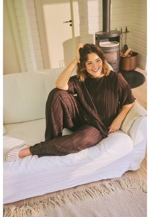 Frau in entspanntem dunklem Outfit sitzt auf weißem Sofa und lächelt, neben einem Holzofen in einem hellen, gemütlichen Wohnzimmer mit hellem Holzboden.