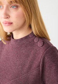 Bordowy sweter z wysokim kołnierzem i trzema ozdobnymi guzikami na ramieniu. Wykonany z miękkiego, teksturowanego materiału z delikatnym melanżem.