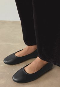 Melnas plakanas kurpes ar apaļu purngalu un zemu sānu daļu, izgatavotas no gludas materiāla, kopā ar tumšām biksēm, novietotas uz gaišas grīdas.