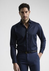 Camisa de vestir azul marino hecha de una tela suave, con cuello abotonado y mangas largas, y un sutil logo bordado en el pecho.