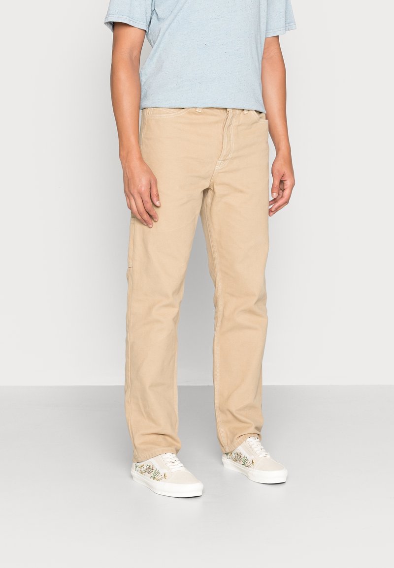 Homme portant un t-shirt bleu clair, un pantalon beige droit et des baskets blanches avec des motifs floraux, debout devant un fond uni.