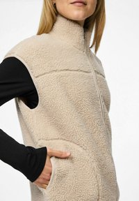 Vrouw in een beige fleece mouwloze vest met rits aan de voorkant en zijzakken, met hand in de zak gestoken, over een zwart longsleeve shirt.