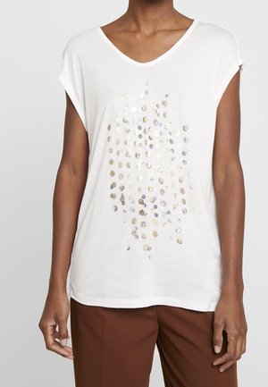 Vrouw draagt een wit T-shirt met korte mouwen en een V-hals met een patroon van zilveren en gouden stippen, gecombineerd met een bruine broek.