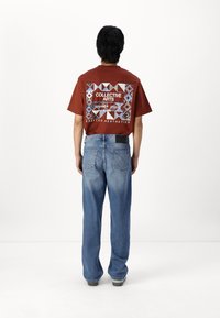 Personne vue de dos portant un t-shirt couleur rouille avec un motif géométrique et le texte "COLLECTIVE ARTS" associé à un jean bleu ample.