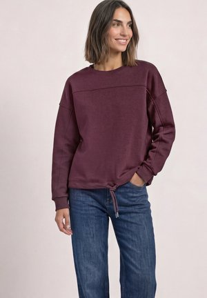 Femme portant un sweat-shirt bordeaux à manches longues et un jean bleu, souriante et debout avec une main dans la poche, devant un fond uni.