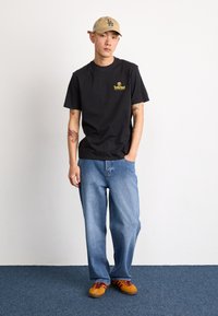 Camiseta negra de manga corta con un logo amarillo de Timberland, combinada con unos vaqueros anchos de color azul claro y unas zapatillas marrones con cordones rojos. Se lleva una gorra beige.