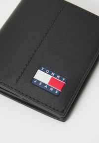 Juodas odinis piniginė su siuvinėtomis detalėmis ir spalvingu "TOMMY JEANS" logotipu, kuriame yra mėlynos, baltos ir raudonos akcentai.