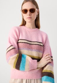 Vaaleanpunainen villaneule, jossa on moniväriset vaaka-raitakuviot beige, ruskea, violetti, vihreä ja sininen. Ominaisuuksiin kuuluu ribbireunainen kaula-aukko ja hihansuut.