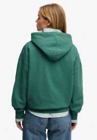 Sudadera verde con capucha, de corte holgado y puños acanalados. Se lleva sobre una camisa de rayas con cuello, visible en el dobladillo. Tela suave y texturizada.