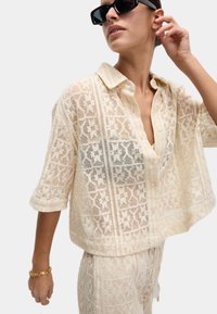 Spitzenstrick, cremefarbige Bluse mit kurzen Ärmeln und Knopfverschluss, ausgestattet mit geometrischen Mustern und einem lockeren Schnitt. Kombiniert mit passenden Hosen.