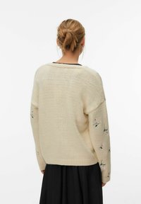 Pleten pulover v nežno beli barvi z ohlapnim krojem, okrašen z rožnim vezenjem na rokavih in rebrastim robom. Gladek tekstura in priložnostna zasnova.