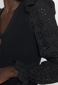 Schwarzes Strickoberteil mit puffärmeln und floralen Ausschnitten. Strukturierter Stoff mit V-Ausschnitt und welligen Ärmelkanten.