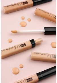 Tubes d'anti-cernes Maybelline Fit Me avec un corps transparent et des caps noirs. Produit beige clair avec un applicateur en forme d'oreille de cerf et des gouttes de liquide beige.